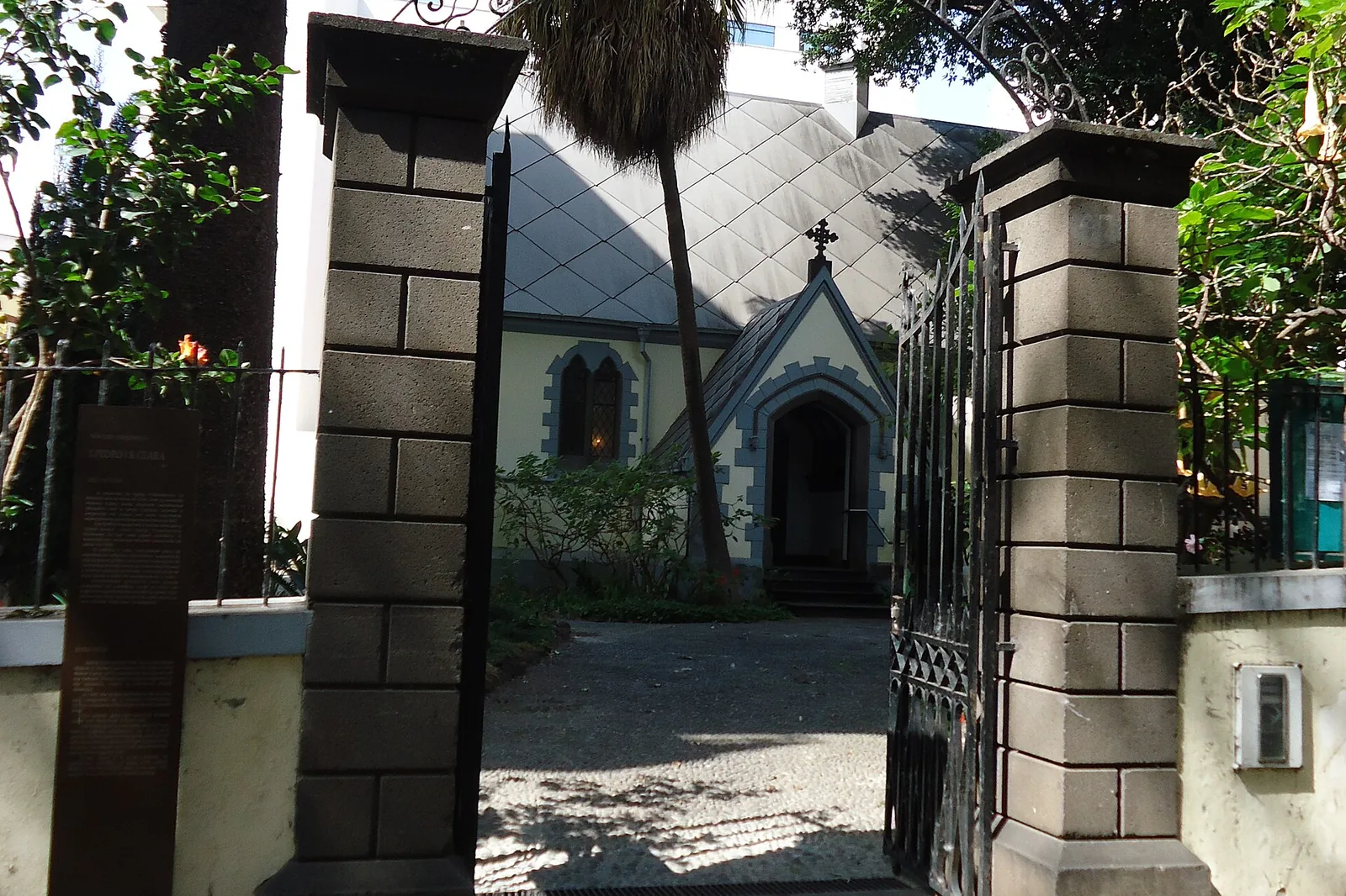 An open iron gate between two stone pillars, with the roof of the church visible in a garden of palms behind it