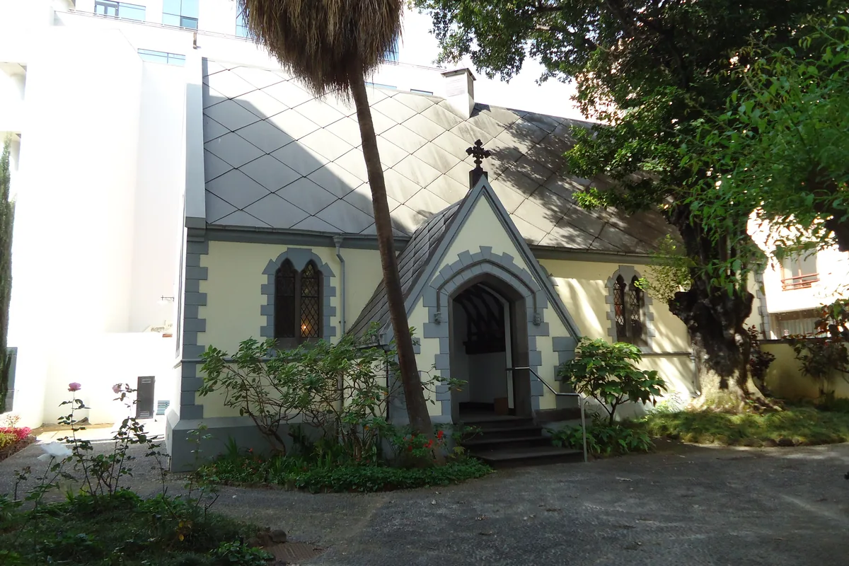 St Andrew's Presbyterian church in Funchal, a small neo-Gothic church with a grey diamond-patterned roof and pale yellow walls, seen across a garden with a leaning palm